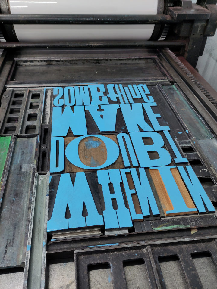 Blue inked wood type forme quoined up in the bed of a large Vandercook proofing press.