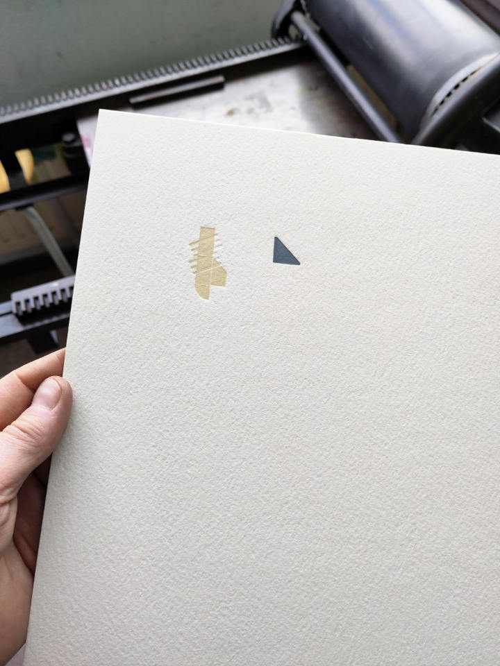 A hand holding a sturdy sheet of white paper. Top left, two small shapes have been printed on it. A gray triangle and a vertical light ochre bar with short diagonal lines. A small proofing press is visible in the background.